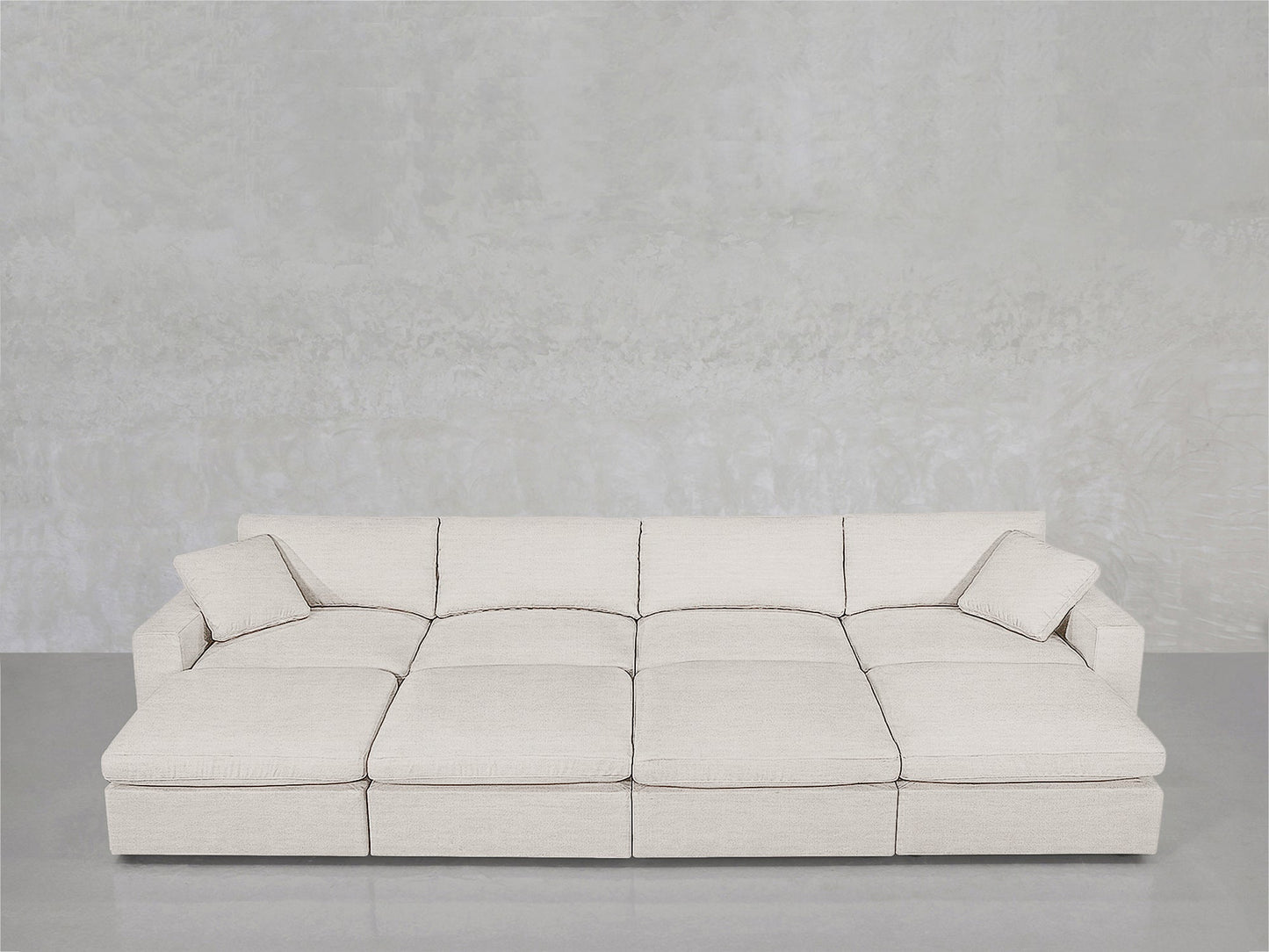 An 8-Seat Modular Daybed in light beige with wide cushions and two matching throw pillows, set against a plain light gray textured wall on a polished floor.