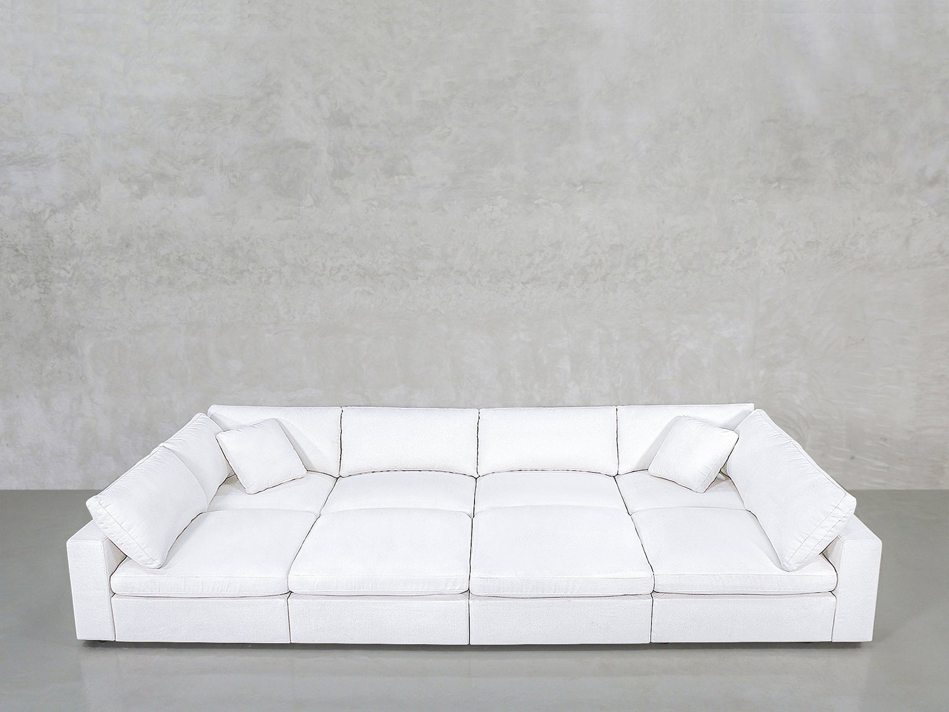 The 8-Seat Modular Pit is a large, modern white sofa featuring wide seats, square armrests, and two matching white cushions, set against a plain light gray wall and smooth concrete floor.