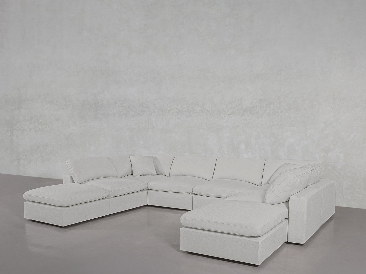 The 8-Seat Modular Double Lounger U-Sectional, large and light gray with multiple cushions, stands on a polished concrete floor against a textured light gray wall.