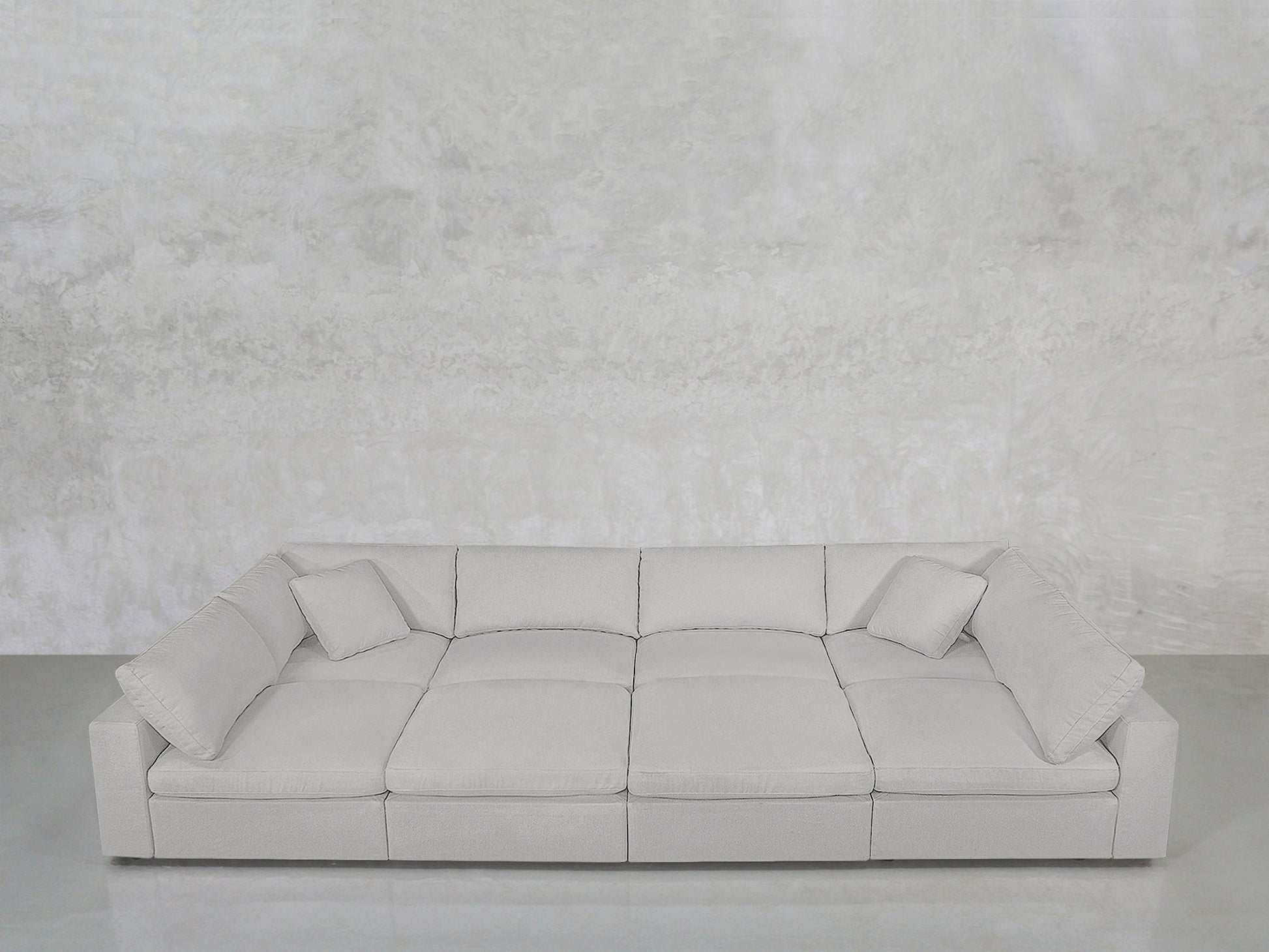 The 8-Seat Modular Pit is a modern, light gray, low-profile sofa with wide armrests and two matching square cushions, shown against a textured light gray wall and smooth gray floor.