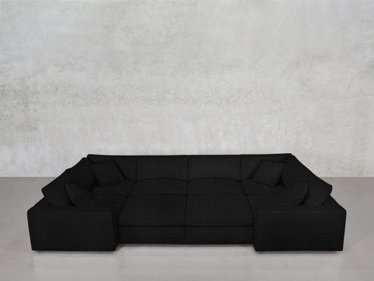 An 8-Seat Modular Closed Pit sectional with four seat cushions and five matching throw pillows sits against a plain, light gray textured wall on a smooth, light floor.