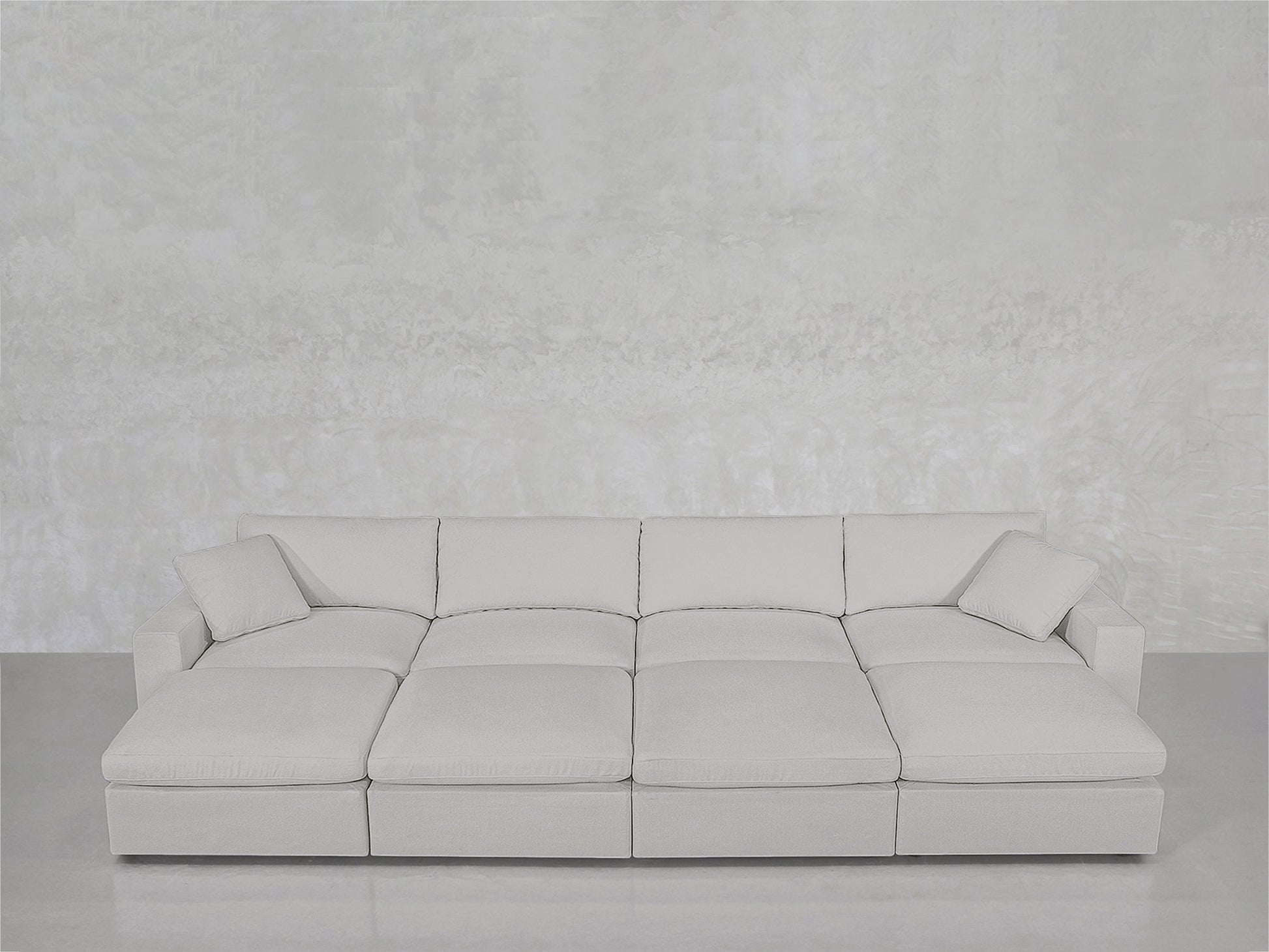 The 8-Seat Modular Daybed in light gray features wide seats and two matching throw pillows, set against a textured pale gray wall atop a smooth, light-colored floor.
