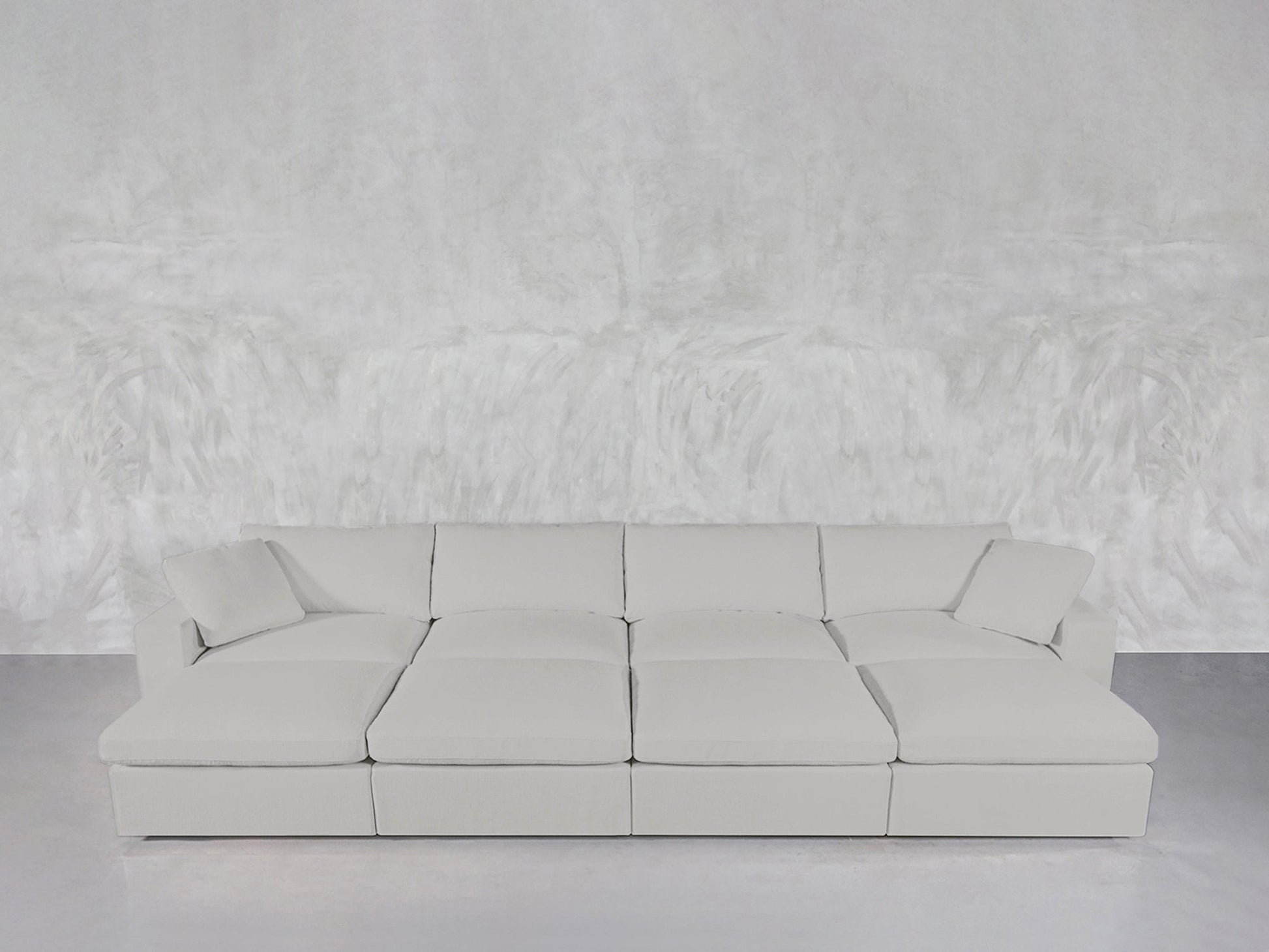 The 8-Seat Modular Daybed, a spacious modern white sectional with wide cushions and four back pillows, sits against a textured white wall on a smooth gray floor.