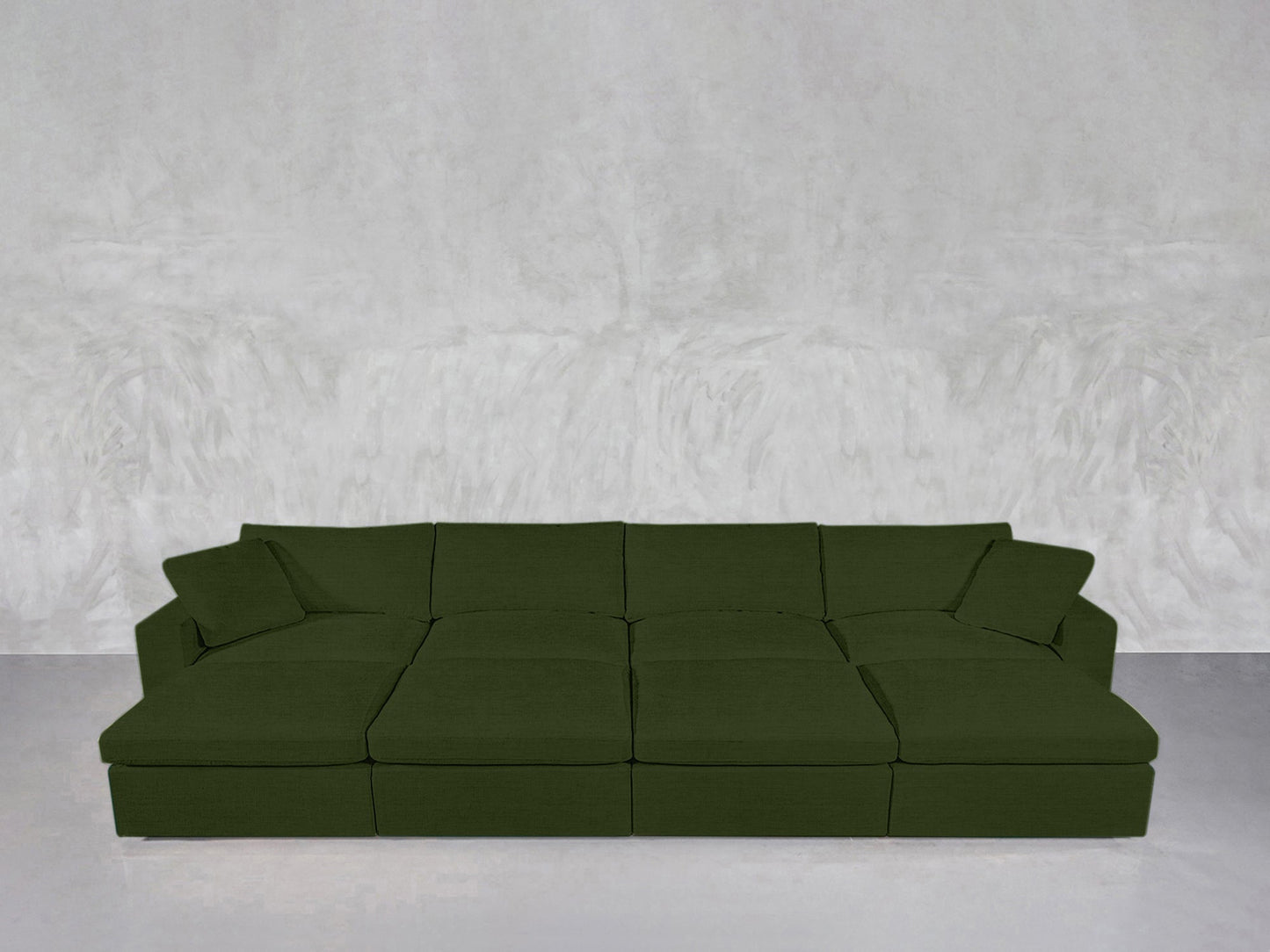 An 8-Seat Modular Daybed in modern green with two matching throw pillows sits against a textured light gray wall on a smooth, light gray floor.
