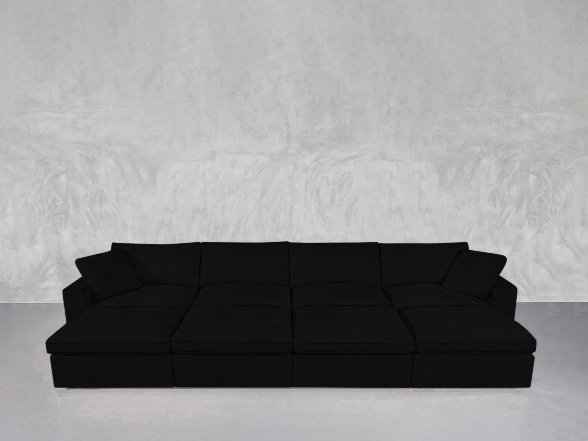 An 8-Seat Modular Daybed in modern black with wide seats and cushions sits against a light gray textured wall and floor in a minimalist room.