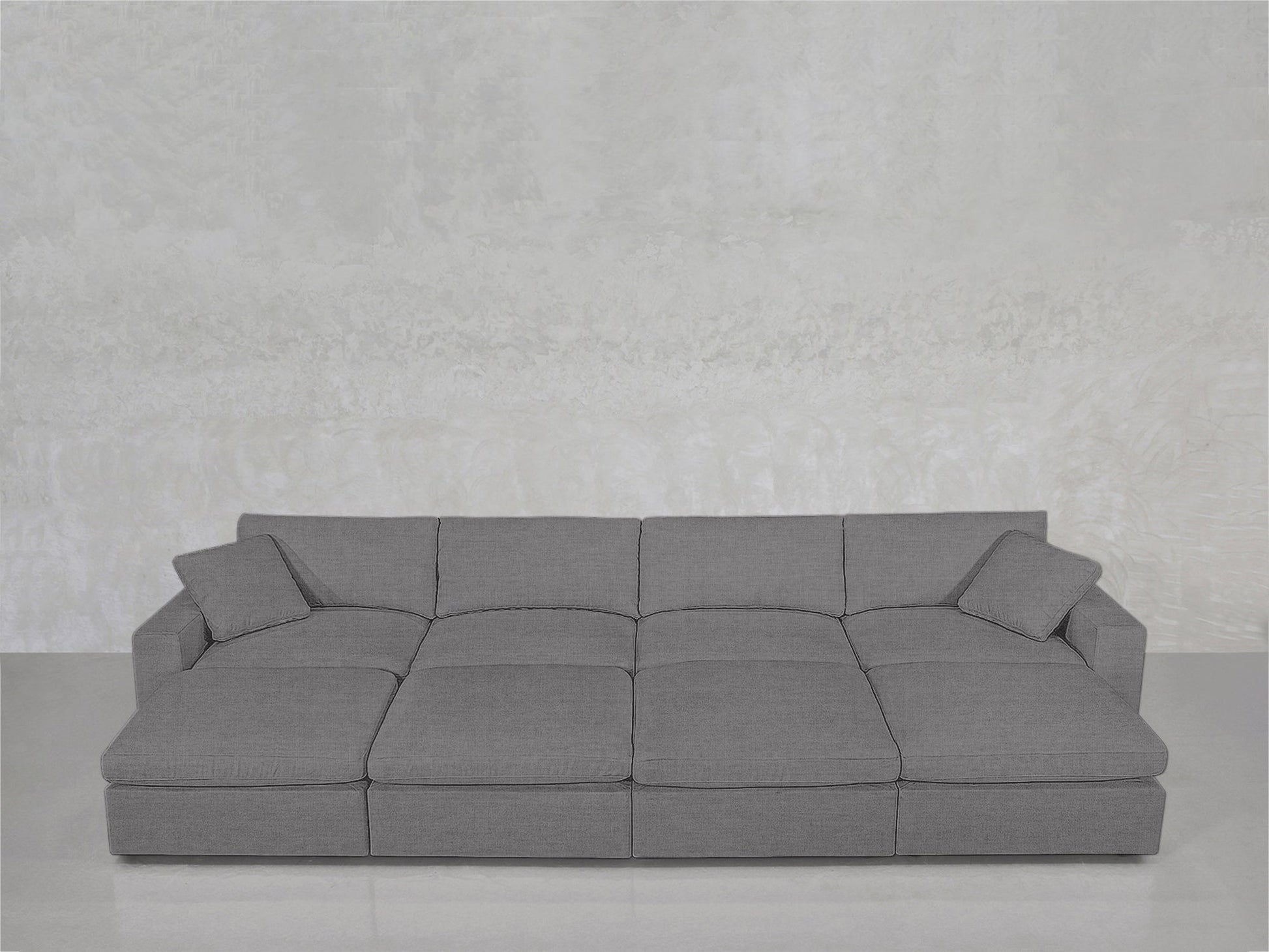 The 8-Seat Modular Daybed, featuring three seat cushions and two matching throw pillows in modern gray, is arranged against a light textured wall with a plain floor.