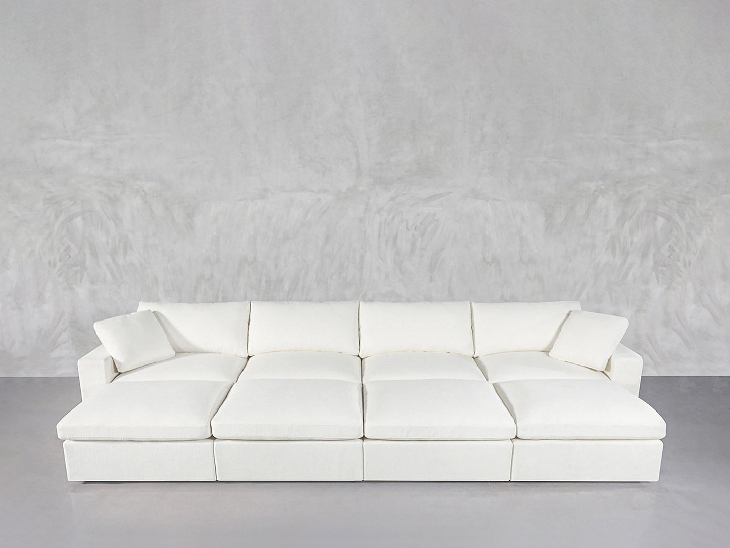 The 8-Seat Modular Daybed in modern white, featuring six large cushions and two matching throw pillows, is arranged against a light gray textured wall and floor.