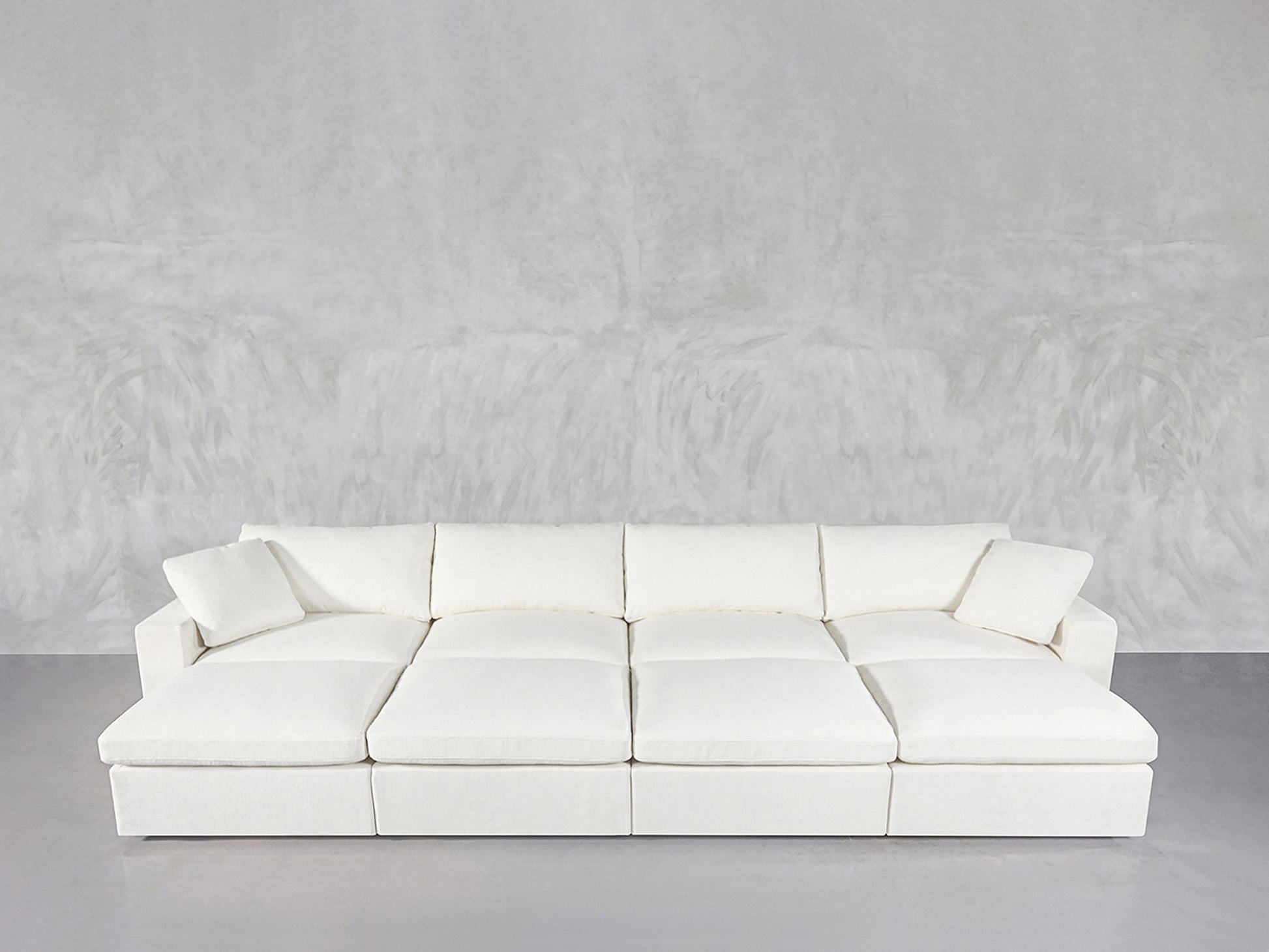 The 8-Seat Modular Daybed in modern white, featuring six large cushions and two matching throw pillows, is arranged against a light gray textured wall and floor.