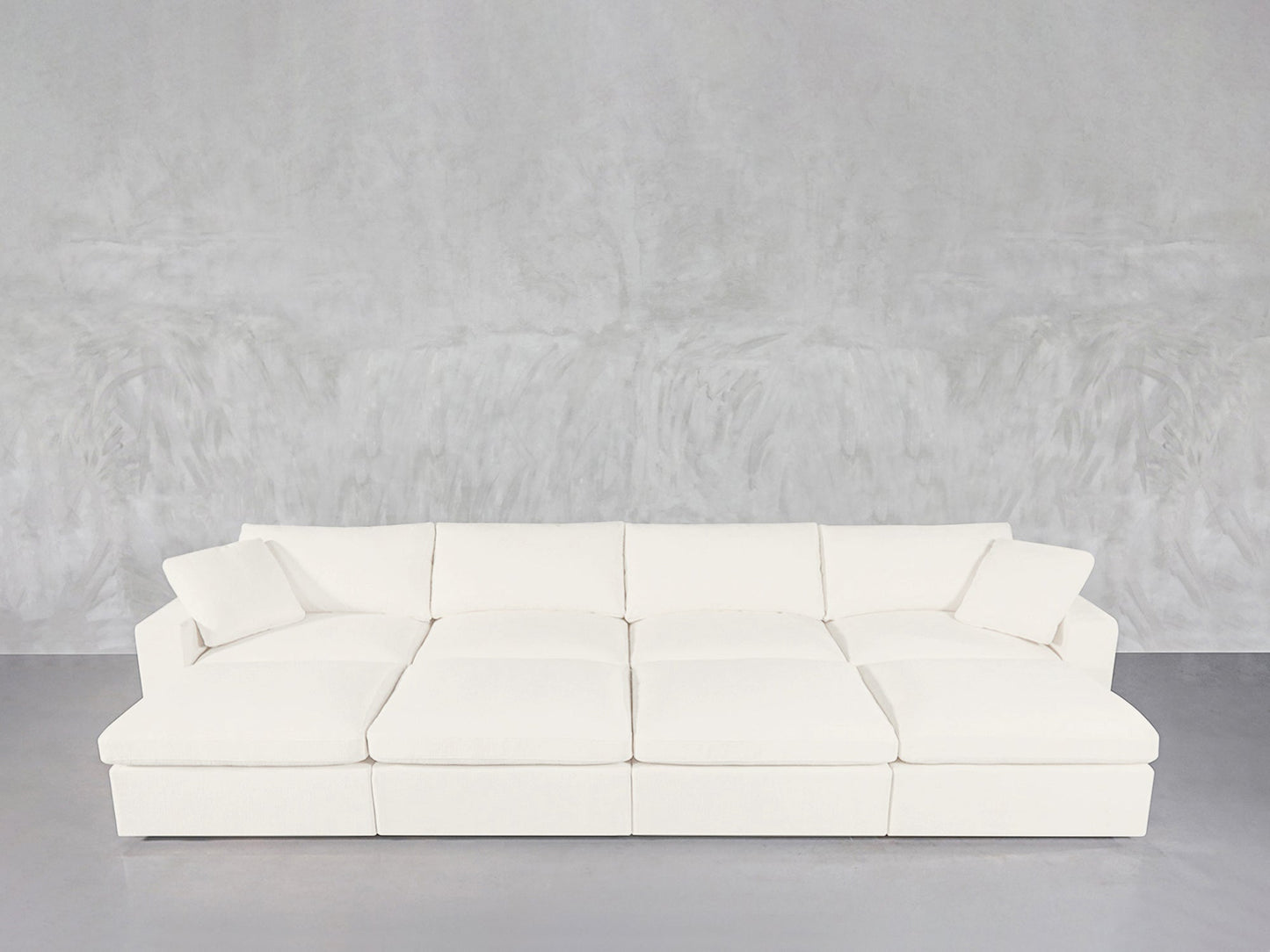 The 8-Seat Modular Daybed is a large, modern white sectional with plush cushions and two matching throw pillows, set against a light gray textured wall and gray floor.