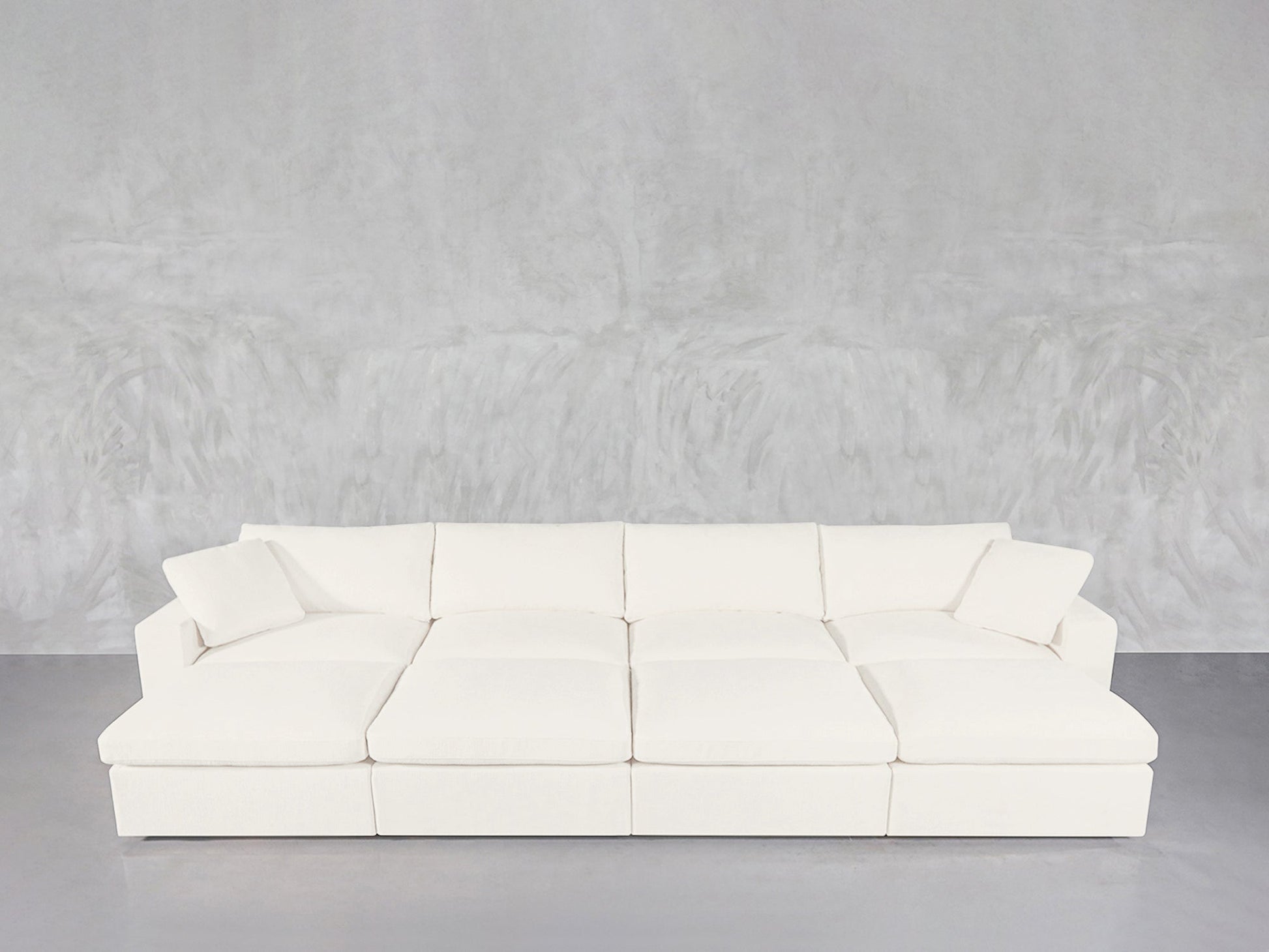 The 8-Seat Modular Daybed is a large, modern white sectional with plush cushions and two matching throw pillows, set against a light gray textured wall and gray floor.