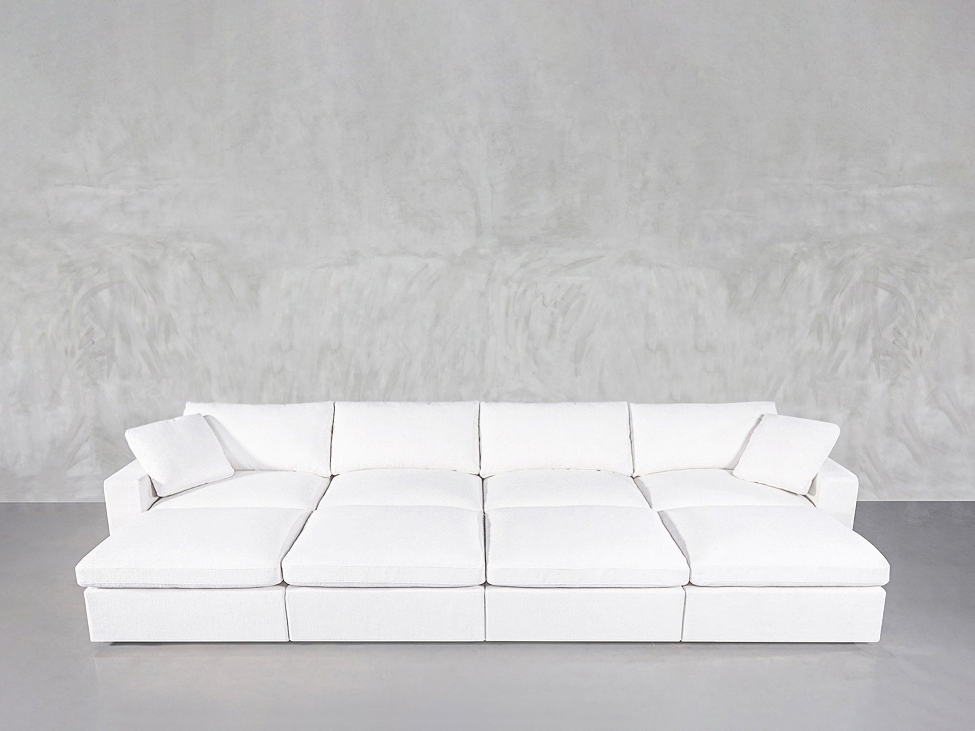 An 8-Seat Modular Daybed with a modern, minimalist design, square cushions, and matching pillows rests against a light gray textured wall on a smooth gray floor.