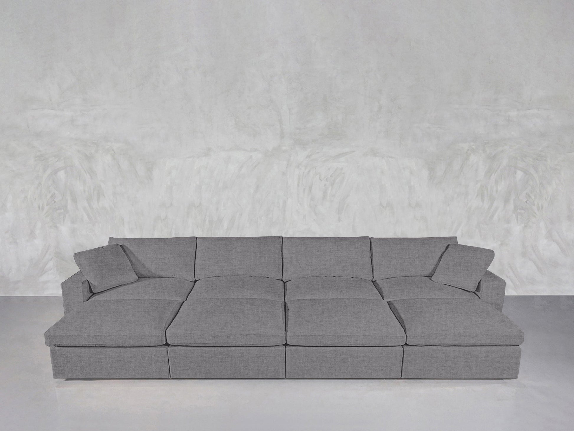 An 8-Seat Modular Daybed in modern gray with three seat cushions and matching throw pillows rests against a textured light gray wall on a polished floor.