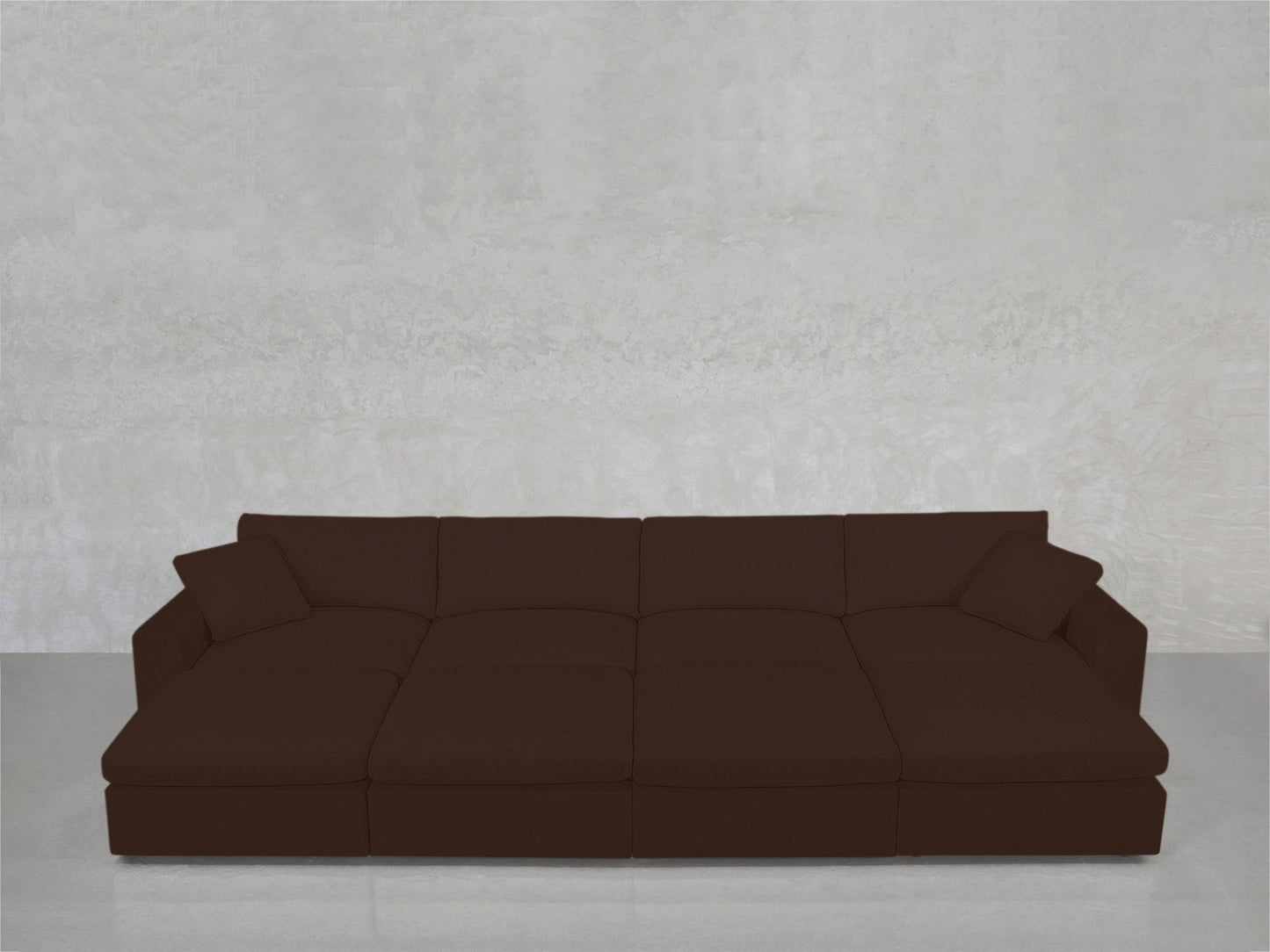 The 8-Seat Modular Daybed, a large modern dark brown sectional with wide seats and two matching pillows, is set against a light gray textured wall and a polished gray floor.