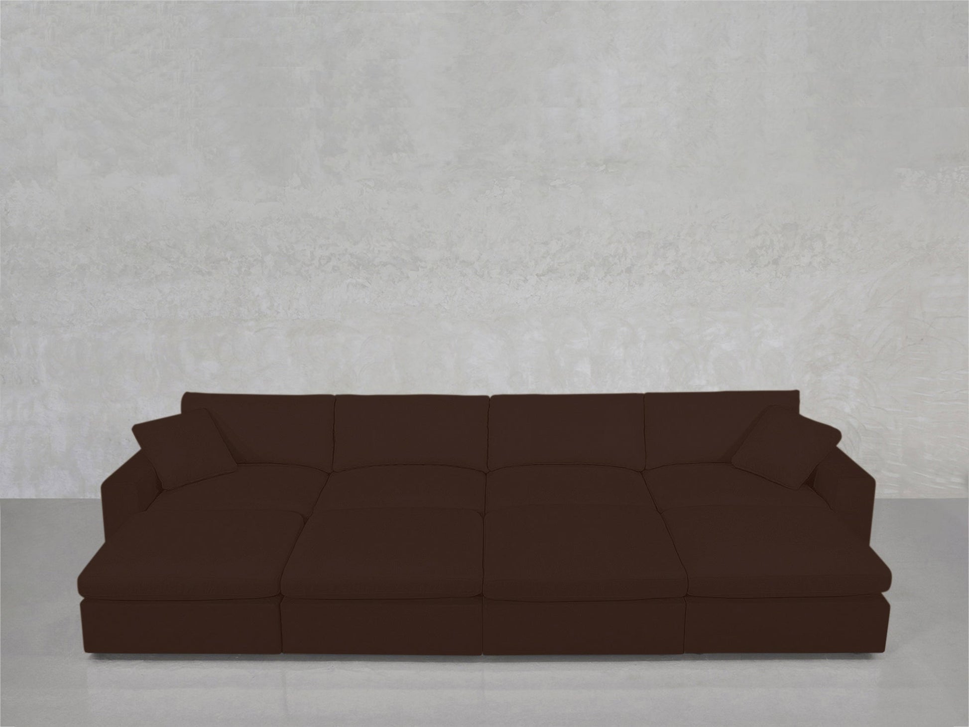 The 8-Seat Modular Daybed, a large modern dark brown sectional with wide seats and two matching pillows, is set against a light gray textured wall and a polished gray floor.