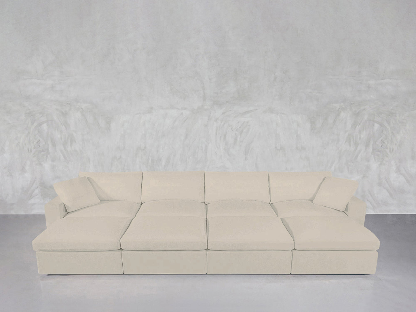 An 8-Seat Modular Daybed in light beige, featuring four seat cushions and four back pillows, is placed against a textured light gray wall and a smooth gray floor.