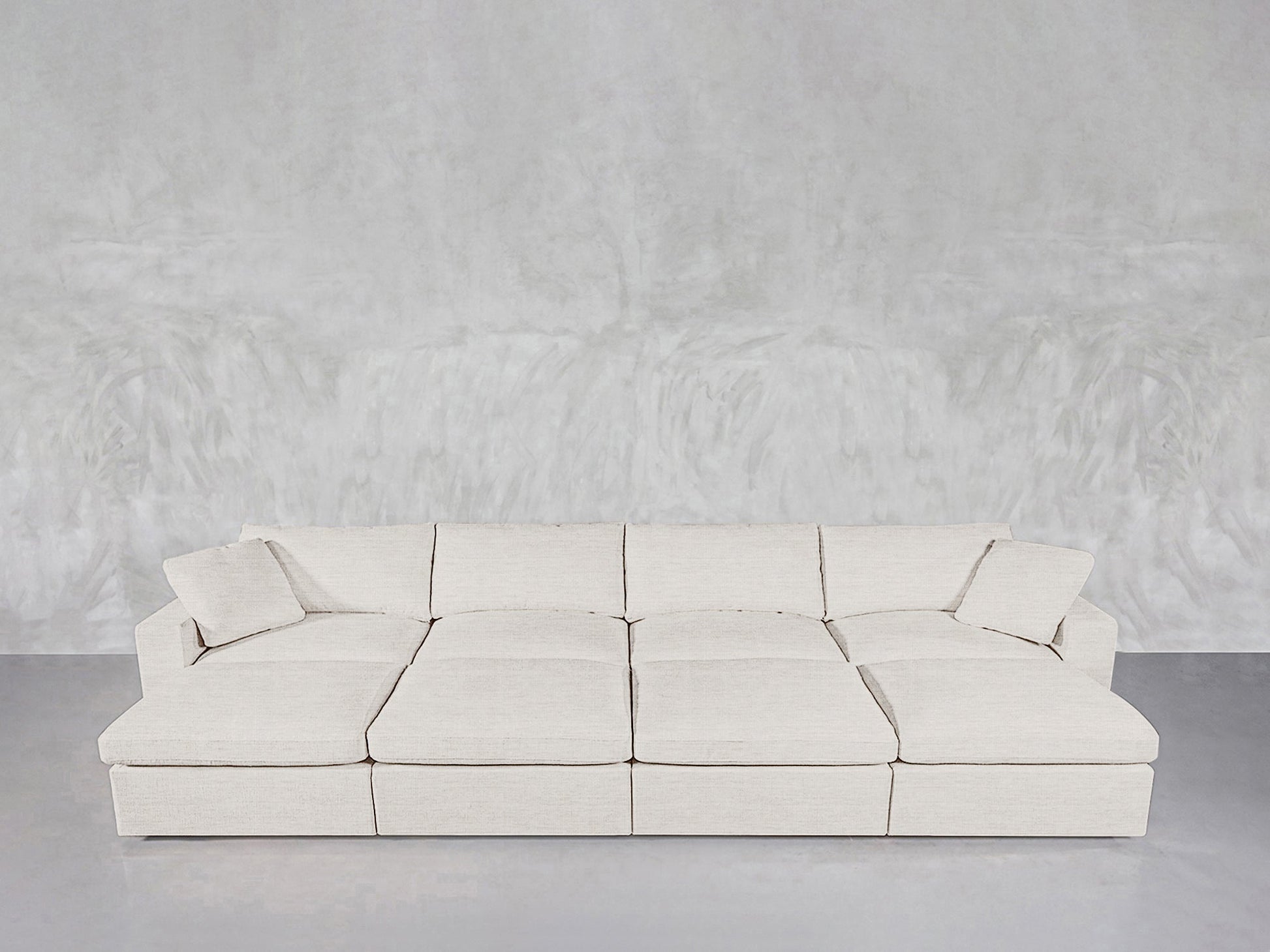 An 8-Seat Modular Daybed in modern white, featuring six seat cushions and two matching throw pillows, is set against a textured light gray wall on a smooth gray floor.