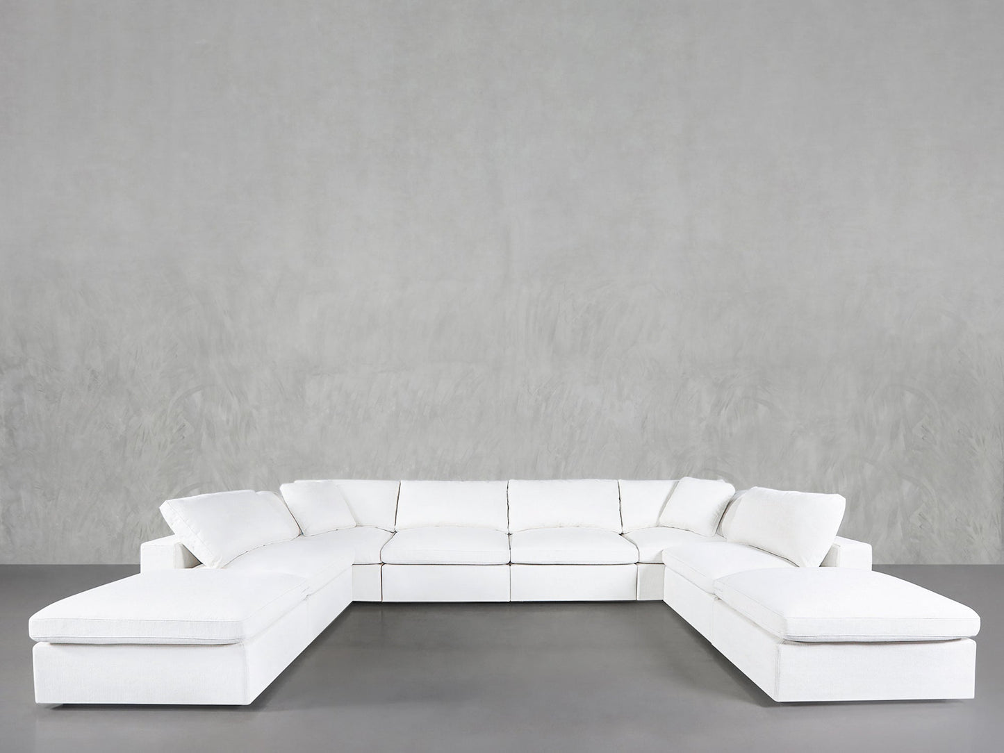 The 8-Seat Modular Double Lounger U-Sectional, in white with plush cushions, is arranged against a plain light gray wall in a minimalist room featuring a smooth gray floor.