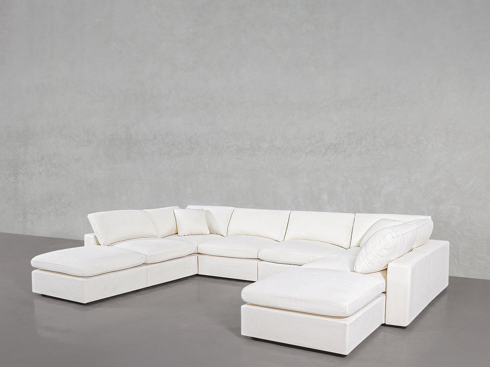 The 8-Seat Modular Double Lounger U-Sectional, featuring plush cushions and chaise lounges at both ends, sits in a minimalistic room with a light gray wall and polished concrete floor.