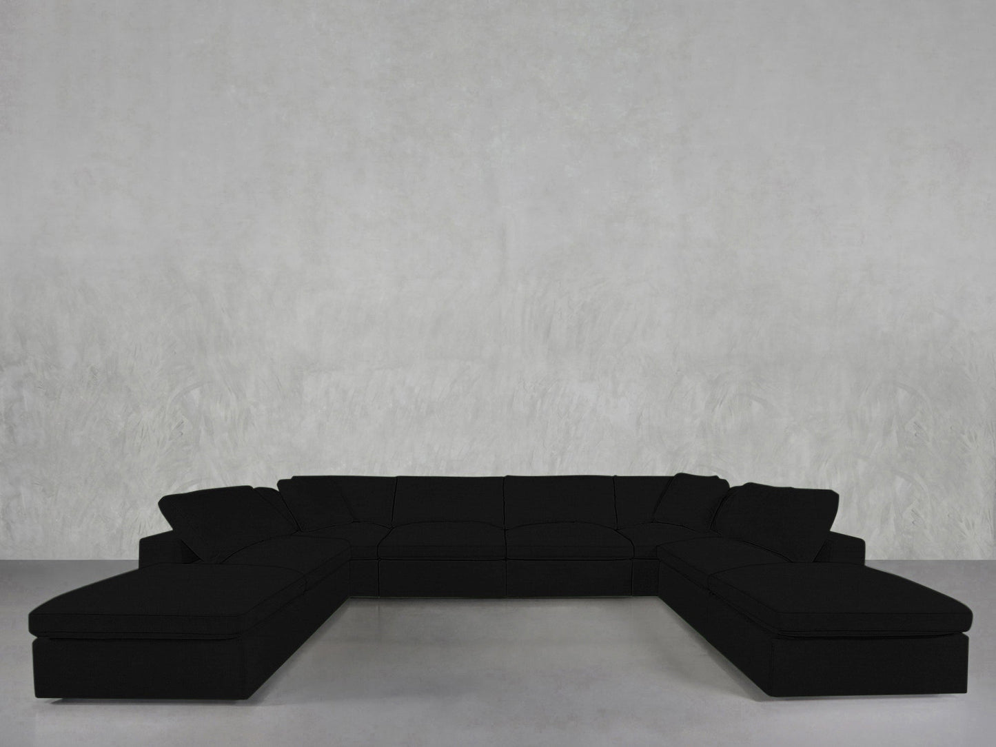 An 8-Seat Modular Double Lounger U-Sectional in black sits against a plain light gray wall on a smooth gray floor, creating a minimalistic setting with no other furniture or decorations visible.