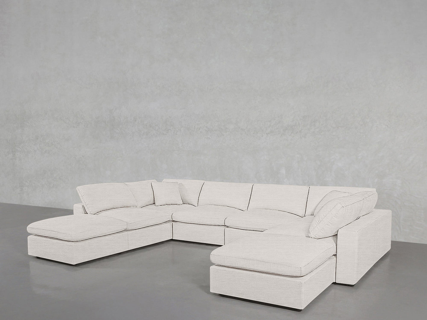 The 8-Seat Modular Double Lounger U-Sectional, a modern light-colored sofa with chaise lounges on both ends, is set in an empty room featuring gray flooring and a textured light gray wall.