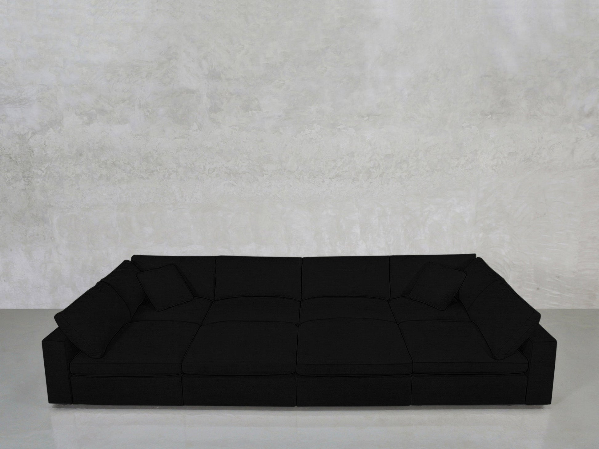 An 8-Seat Modular Pit in modern black with low backrests and three cushions sits against a light gray textured wall on a smooth, light-colored floor.