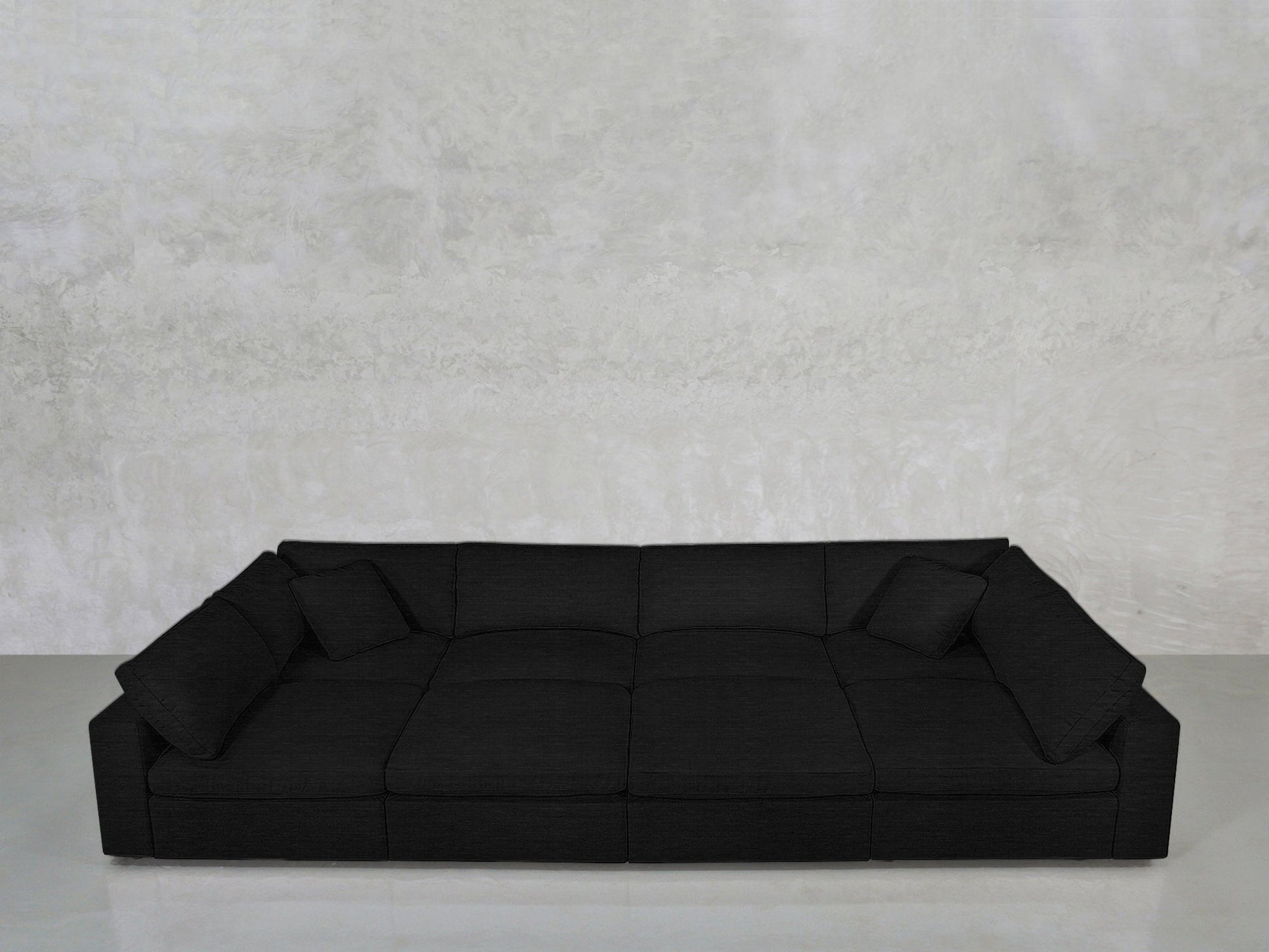 The 8-Seat Modular Pit is a modern, low-profile black sofa with wide seats and two matching black cushions, set against a light gray textured wall on a smooth, light-colored floor.