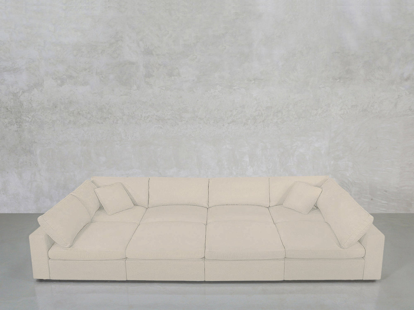 The 8-Seat Modular Pit sectional in light beige, with four matching cushions, is set against a textured gray wall and rests on a smooth gray floor.