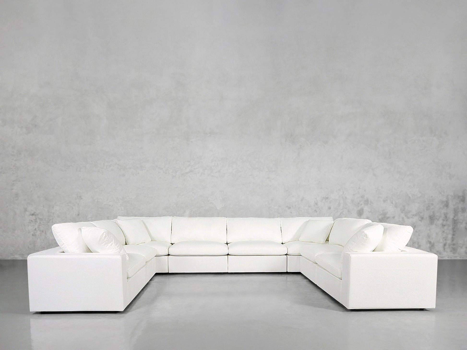 An 8-Seat Modular U-Sectional with matching cushions is arranged against a plain, light gray wall and floor in a minimalist, modern room.