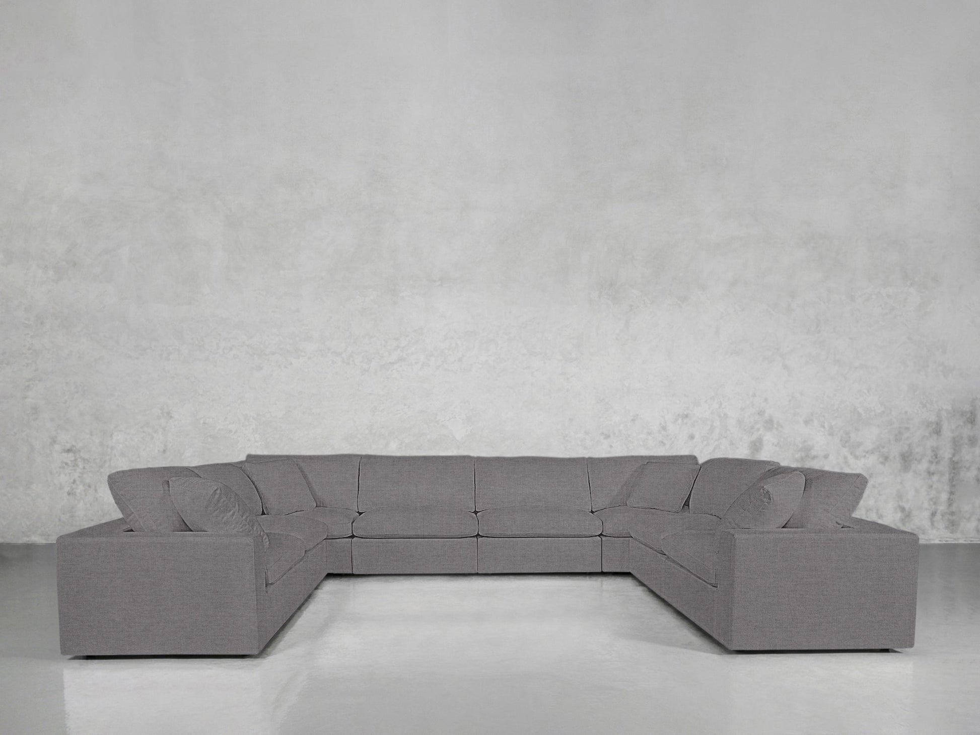 An 8-Seat Modular U-Sectional with a modern gray design and matching cushions is set in a minimalist room with a light gray textured wall and a sleek, shiny floor.