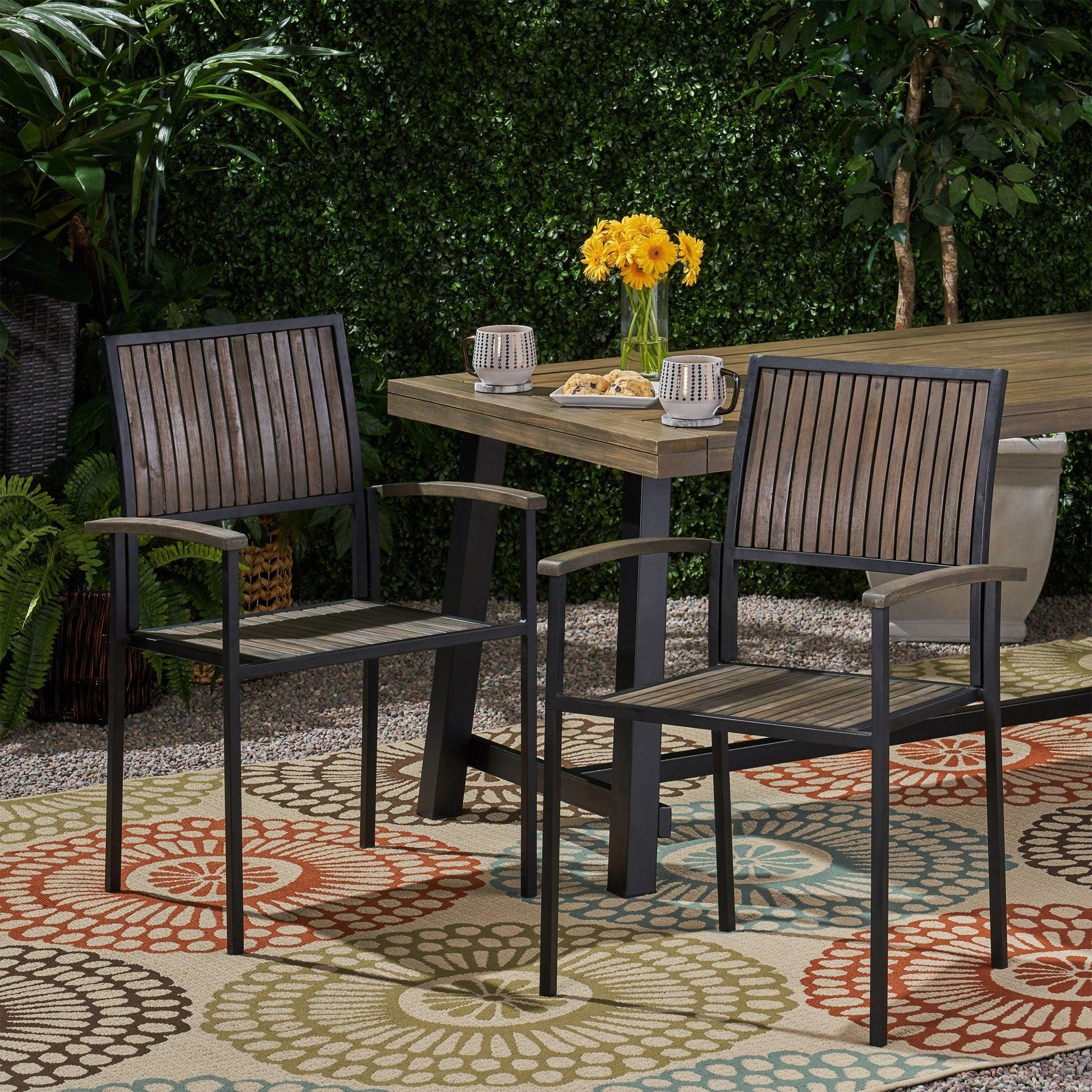 Two Vermilion Outdoor Dining Chairs with vertical slats and wooden arms sit on a colorful rug by a rectangular table topped with yellow flowers in a garden surrounded by greenery.