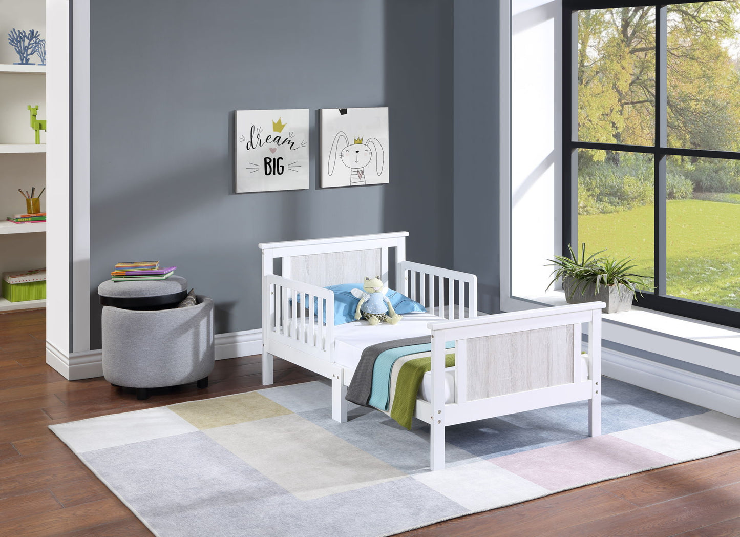 A child's bedroom features the Connelly Reversible Panel Toddler Bed in white with striped bedding, a stuffed bunny, and gray walls displaying two framed prints. Sunlight streams through a large window. A gray ottoman and bookshelves sit in the corner.