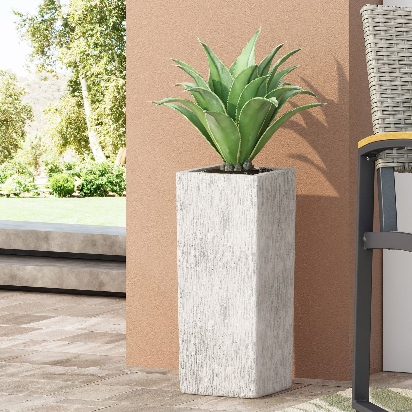 The Modern Outdoor Planter, a tall white rectangle holding a green succulent, sits on a tiled patio beside a beige wall and wicker chair, with trees and grass in the background.
