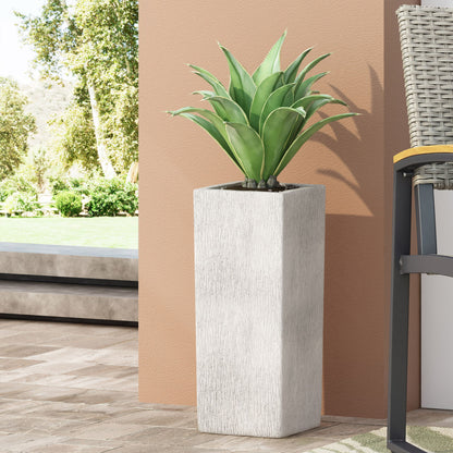 The Modern Outdoor Planter, a tall white rectangle holding a green succulent, sits on a tiled patio beside a beige wall and wicker chair, with trees and grass in the background.