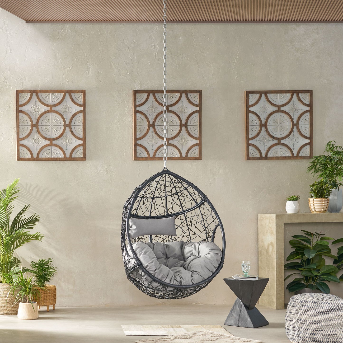 A modern room features the Teardrop - Hanging Chair With 8Ft Chain and gray cushion, surrounded by green plants, a small triangular side table with a glass, and three framed wall art pieces. Neutral tones dominate the space.