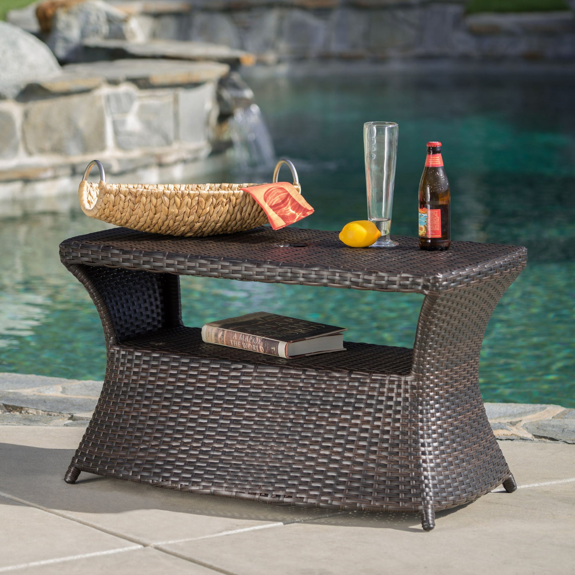 The Berkeley - Chaise Lounge Side Table sits by the pool, holding a straw basket with chips, lemon, an empty glass, and a bottled drink. A book rests on its lower shelf, with stone decking and water in the background.