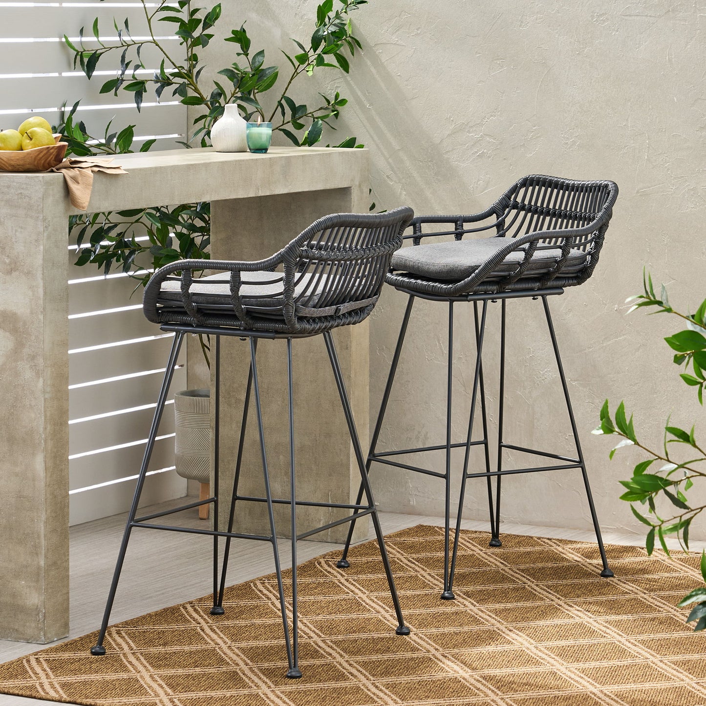 Two Outdoor Rattan Bar Chairs with metal legs and gray cushions stand by a concrete bar on a modern patio, complemented by green plants and a tan patterned rug.