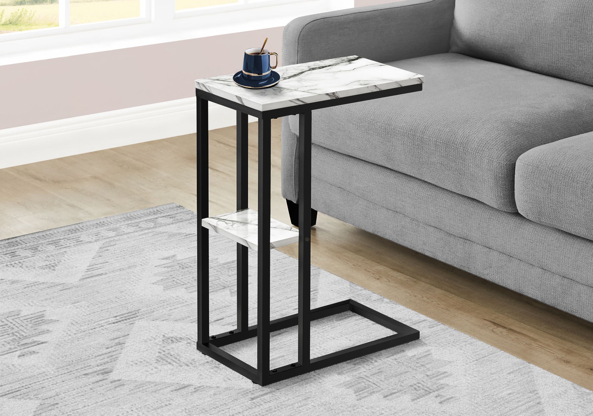 The Accent Table C-Shaped, Contemporary & Modern with a marble top and black metal frame stands by a gray sofa. On the table is a blue cup and saucer with a spoon. The room has a patterned rug and wooden floor.