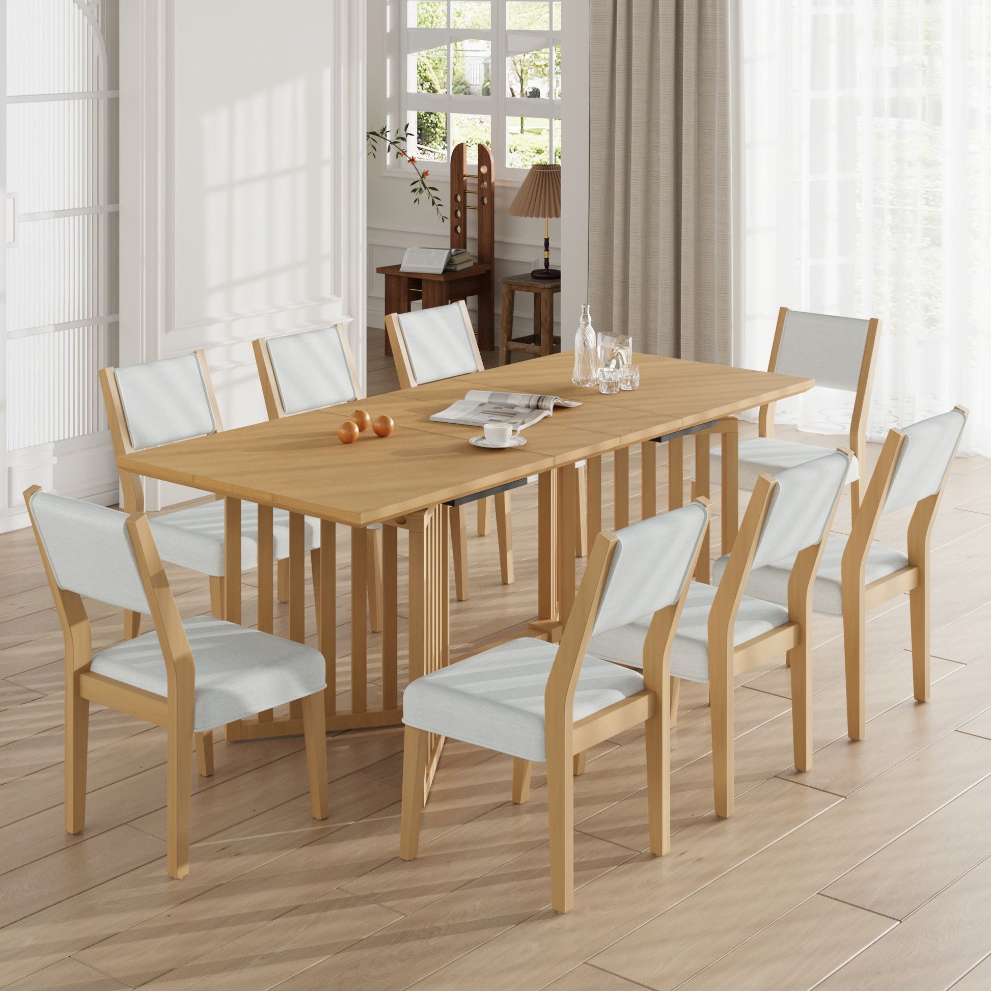 A modern dining room featuring the Topmax 9 Piece Farmhouse Extendable Dining Table Set with two removable leaves and eight upholstered chairs, styled with a glass pitcher, glasses, and a bowl of oranges on a light wood floor near large windows.