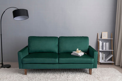 A Comfortable Loveseat With Durable Construction sits on a white rug in a minimalist room, flanked by a black floor lamp on the left and a small shelf with books and framed art on the right, all set against a gray wall.