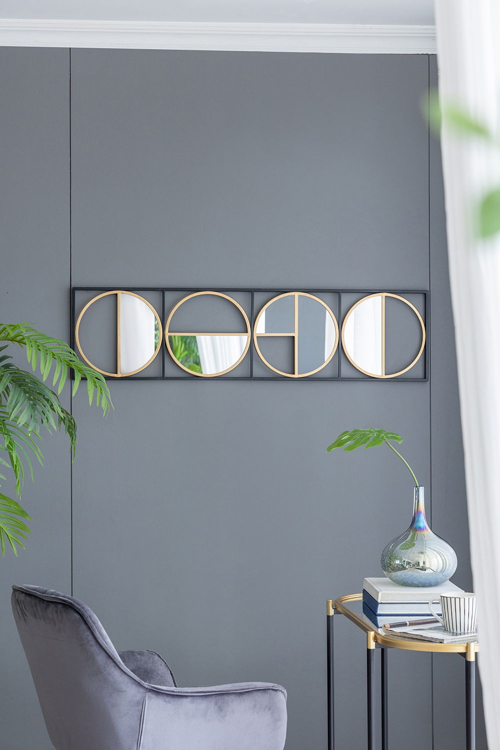 A modern living room corner with a gray wall, an Eclectic Styling Metal Beaded Wall Mirror, a gray velvet chair, a small gold side table with a glass vase, a green plant, and natural light streaming through sheer white curtains.