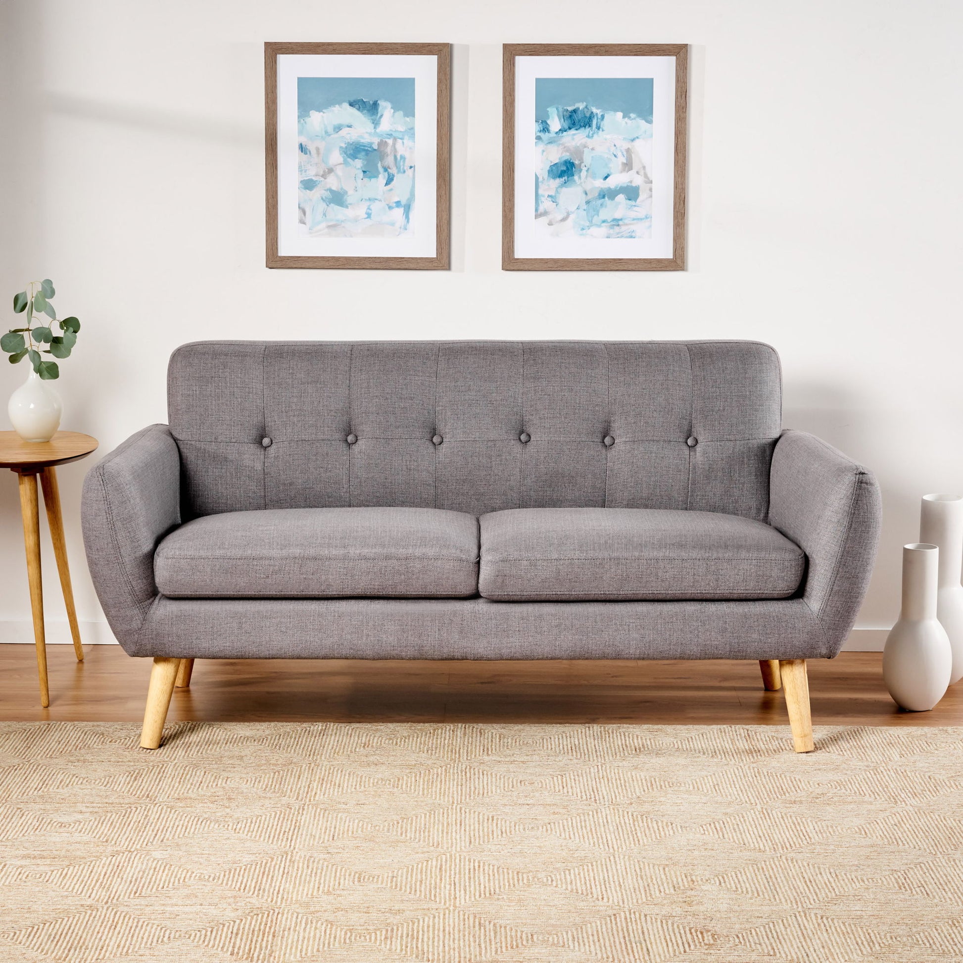 A Modern Loveseat Sofa With Button Tufted Backrest sits against a white wall, flanked by a round side table with a plant and white vases. Two abstract blue and white artworks hang above the sofa.