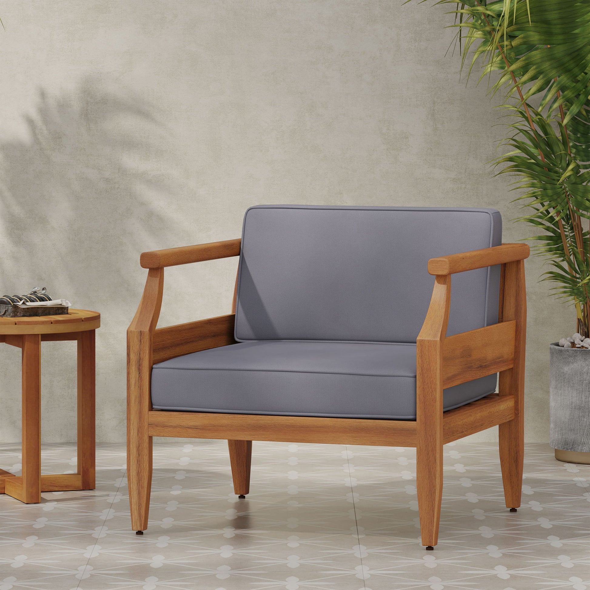 An Outdoor Club Chair With Recessed Arms And Cushions in gray sits on a tiled floor next to a small round wooden table, with two potted plants against a light-colored wall in the background.