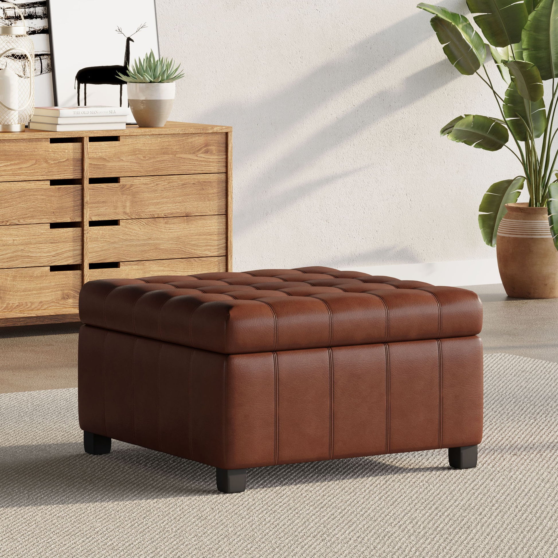 A Storage Ottoman For Living Room, featuring tufted brown leather, sits on a light rug in a modern space with a wooden dresser, potted plant, and decorative accents in the background.