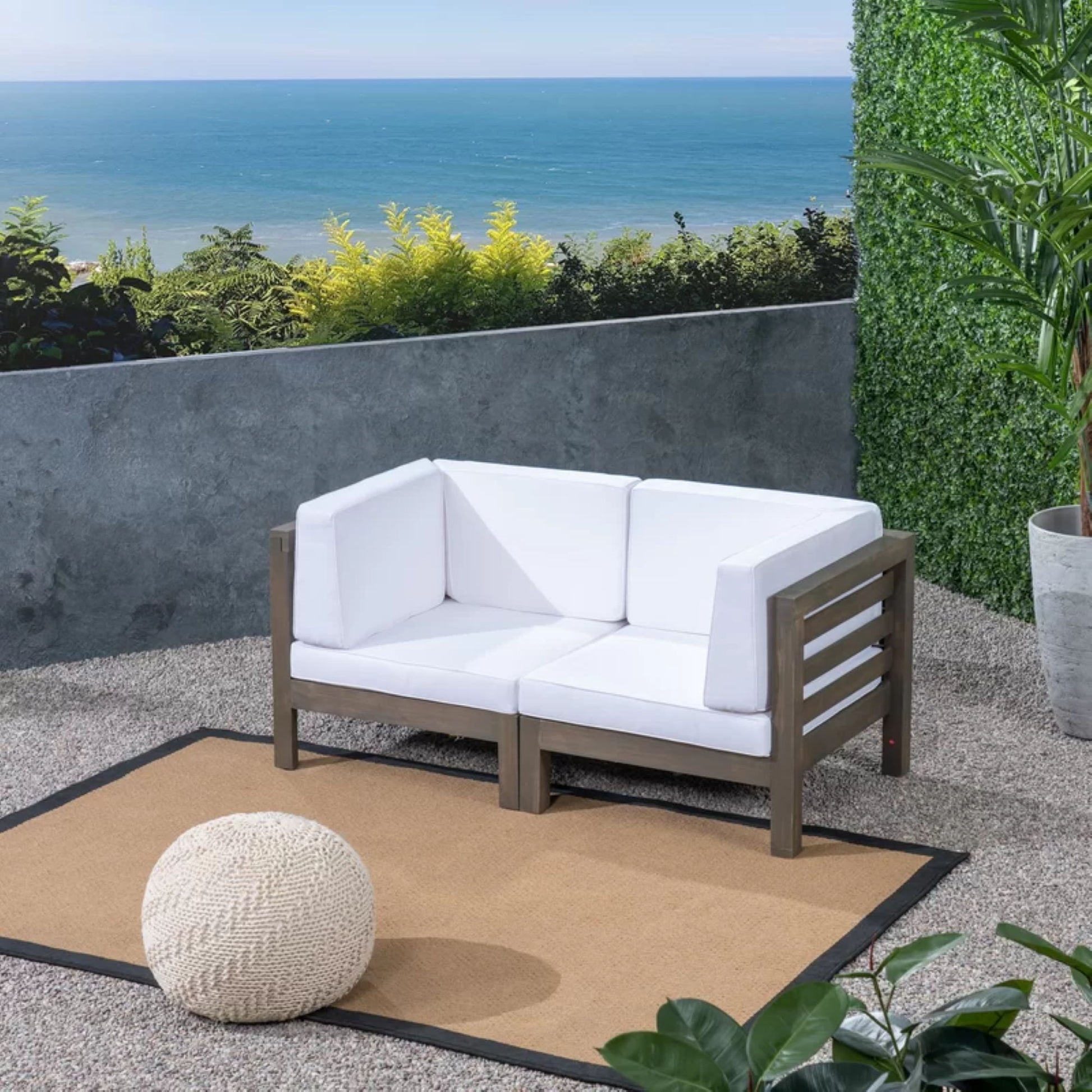 Modern outdoor seating area featuring the Oana 2 Piece Loveseat Set with water resistant cushions, arranged on a tan rug beside a knitted pouf and green plants, overlooking a scenic ocean view under a blue sky.