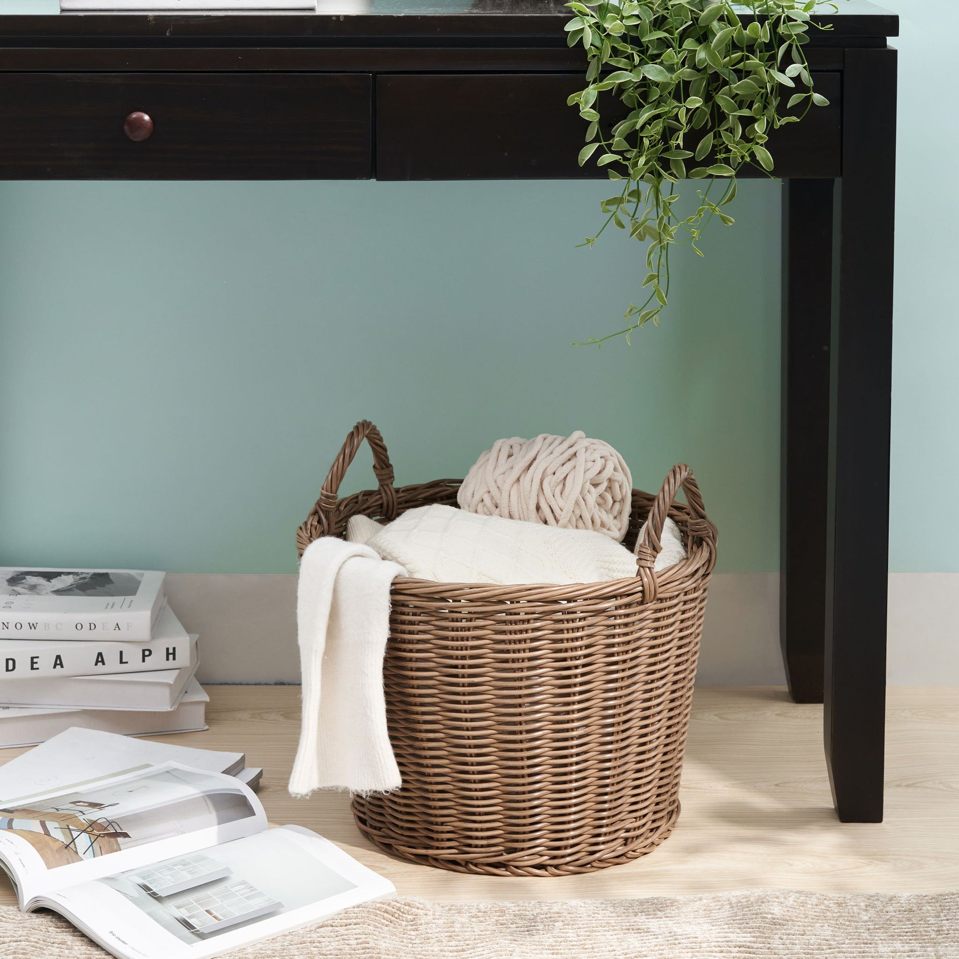 A Round Cone Woven Resin Wicker Basket With Handles in walnut brown, filled with a white blanket and knitted throw, sits under a dark wooden desk with a small plant. Books and magazines are scattered on the light wood floor nearby.