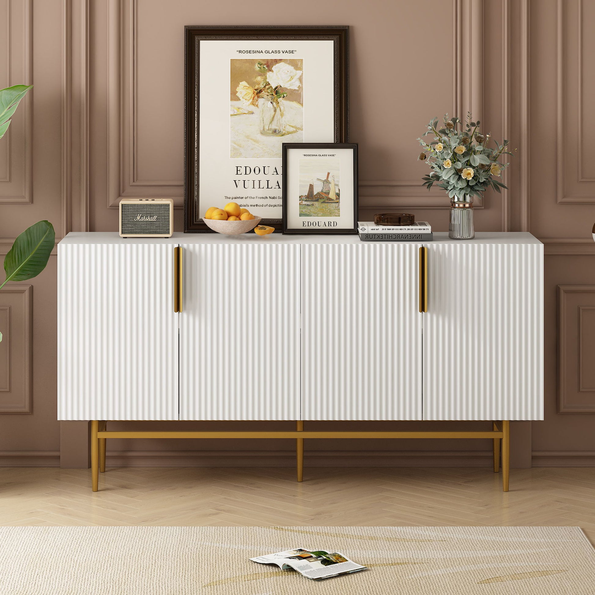 The Modern Elegant 4 Door Sideboard Gold Metal Handle Buffet Cabinet stands against a brown paneled wall, topped with framed art, flowers, a bowl of oranges, and a book; an open magazine lies on the floor beside it.