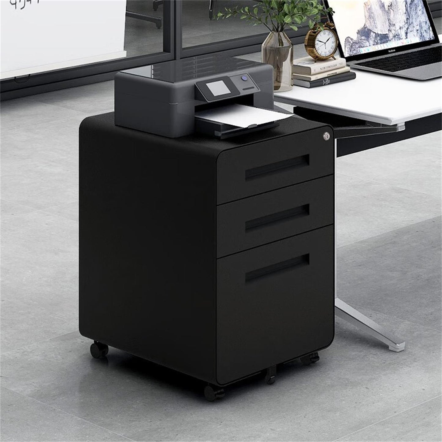 A 3 Drawer Mobile File Cabinet with a 5-wheel anti-tilting design, made of cold rolled steel, sits under a desk in a bright office. A printer on top prints paper beside a modern desk with a laptop and plant. Suitable for Legal/Letter/A4 files.