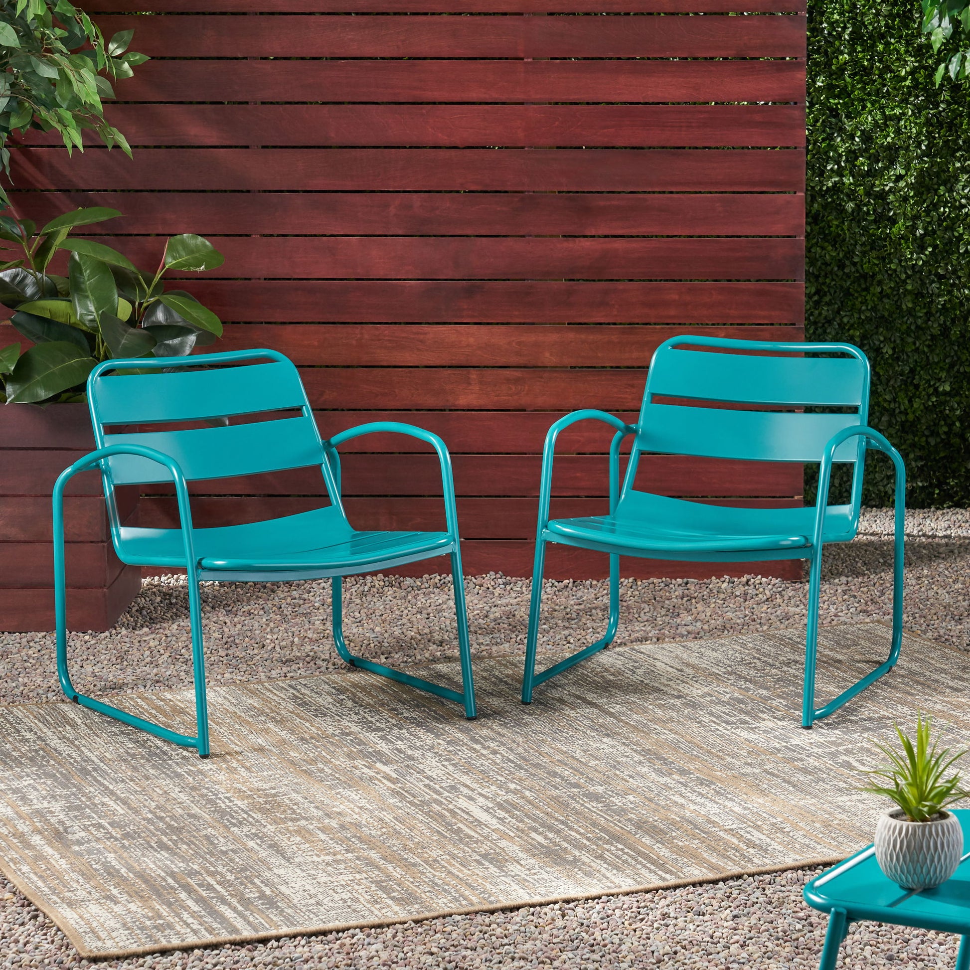 Two Conrad teal metal outdoor chairs are placed side by side on a rug over gravel, set against a wooden slat fence and green plants. A small table with a plant is partially visible in the foreground.