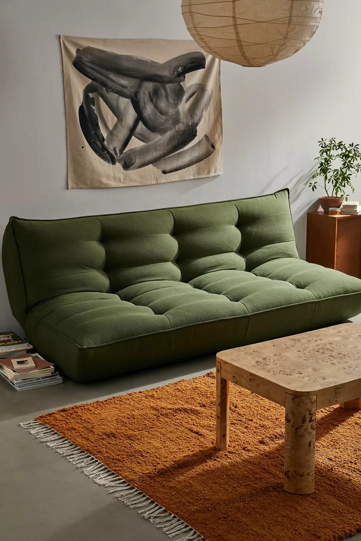 A cozy living room showcases a plush Greta XL Sleeper Sofa with tufted cushions, complemented by a wooden coffee table on an orange fringed rug and a potted plant. A monochrome abstract artwork decorates the light-colored wall, while a hanging spherical light provides soft illumination.