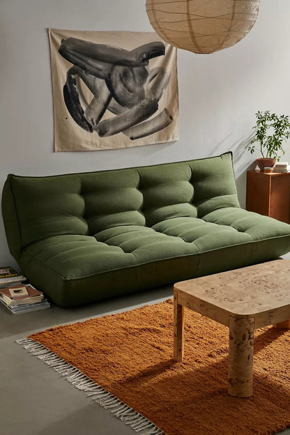 A cozy living room showcases a plush Greta XL Sleeper Sofa with tufted cushions, complemented by a wooden coffee table on an orange fringed rug and a potted plant. A monochrome abstract artwork decorates the light-colored wall, while a hanging spherical light provides soft illumination.